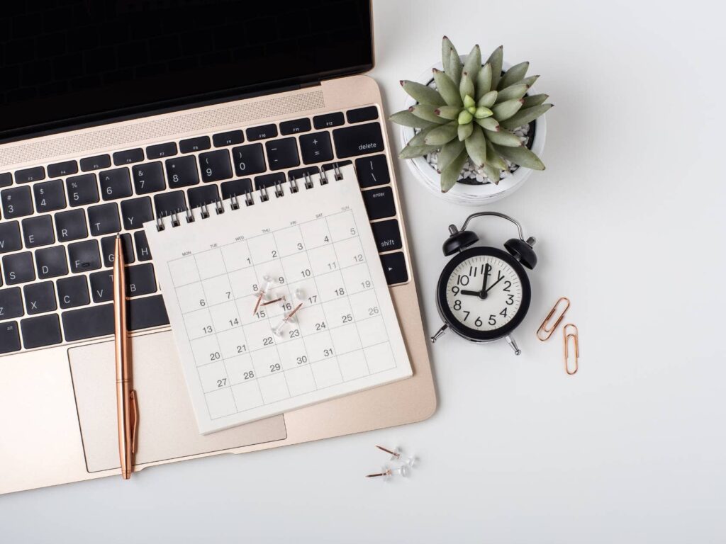 Un calendrier posé sur un clavier d'ordinateur portable avec des épingles marquant certaines dates, accompagné d'un stylo, d'une horloge. Tester différents moments pour envoyer sa newsletter permet d'optimiser son taux d'ouverture et d'engagement.